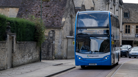 A blue bus with 'Cirencester' as the destination on it drives through a single laned road next to a quaint looking brick wall.