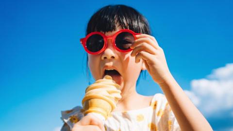 Young girl with dark hair wearing red sunglasses and eating ice-cream cone outdoors on a sunny summer day against blue sky