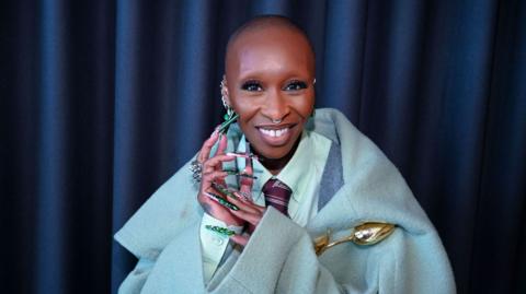 Cynthia Erivo smiles wearing a light blue coat and showing her long green nails