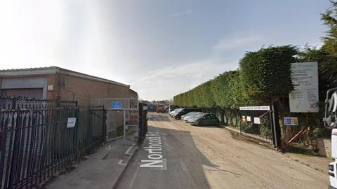 A road next to industrial buildings, with gates leading down the road. Hedges are on the right-hand side of the road and a row of cars are parked further down the road.
