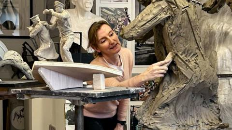 Amy Goodman is pictured in her workshop, using a tool on a statue in progress. Near her are figurines of two men on the prow of a boat, showing how part of the Coastal Forces memorial will look. Photographs of Royal Navy Wrens are positioned in front of her.