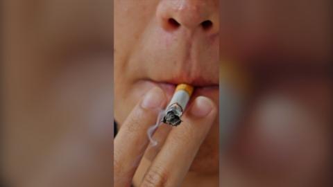 A close-up of a person's face showing a smoking cigarette in their mouth, and two fingers either side of it.