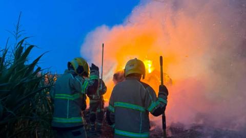 It is evening and the sky is dark blue. Several firefighters wearing protective clothing and yellow helmets are standing near a large farm vehicle engulfed in bright orange flames, with smoke billowing from it, and sparks flying in the air