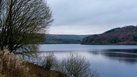 Scammonden Reservoir is shown, a large body of water surrounded by rolling hills in shades of green and brown. There are bare trees and bushes in the foreground, and the sky is wintry and overcast.