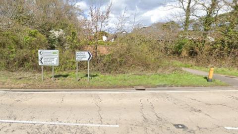 A section of a road with signs for various places, one of which is Lymington. Behind the signs, there is greenery and houses. It's a sunny day.