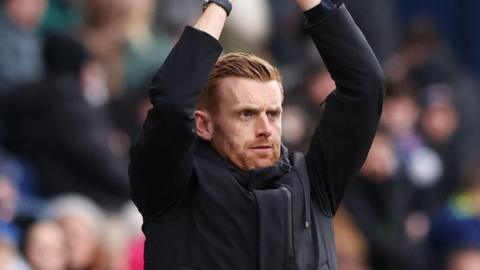 Ed Still standing in the dugout with his arms above his head as he claps his hands