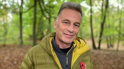 Chris Packham smiles at the camera in Wytham Great Wood. He wears an olive green puffa jacket with a Royal British Legion poppy attached.