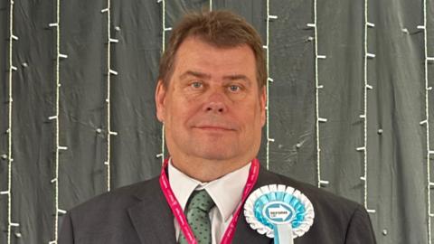 A clean-shaven man with short brown hair is weaing a black suit, pale green dotted tie, a pink lanyard and blue and white Reform ribbon badge