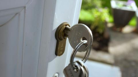 A set of silver keys in a golden lock, attached to a white plastic door. The background is blurry, but grass and a plant pot is visible to the right of the image.