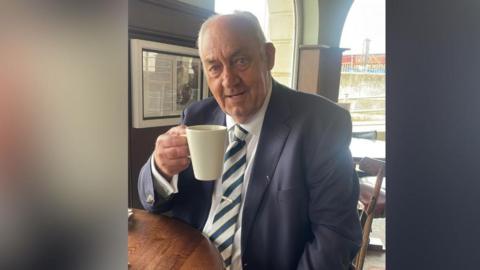 A man with grey hair wearing a blazer, tie and white shirt sitting at a table while holding up a coffee mug.