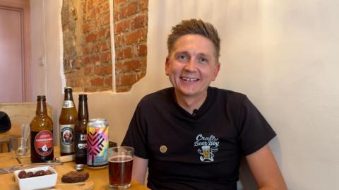Image shows Ryan smiling at the camera. He is wearing a black short-sleeved T-Shirt which says 'Crafty Beer Boy' on it. Behind him is a white wall with exposed brick. Beside him is a table with a selection of beers on it and food which he is pairing together.
