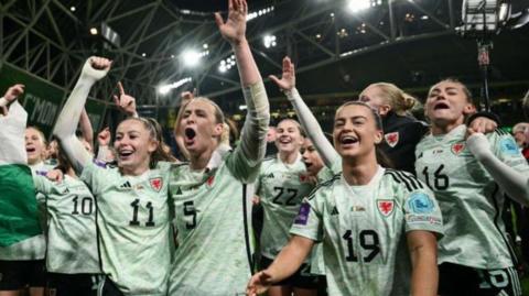 Wales women celebrate win, in their muddied kit with hands in the air