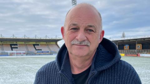 Ian Darler is in the middle. He has a grey moustache and is wearing a blue jumper and a black T-shirt. He is stood on a football pitch which has a light layer of snow covering it.