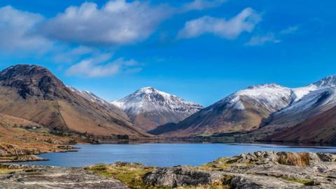 A lake or tarn backed by mountains covered with a light dusting of snow. The sky is bright blue, which is reflected in the lake, with just few high clouds.