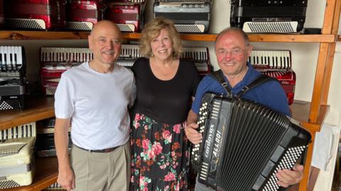 A man, a woman and a man with an accordion smile while standing in front of shelves filled with other accoordions