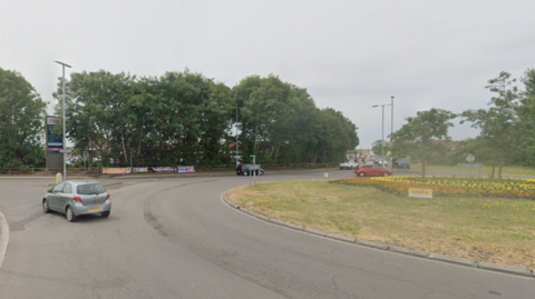 The roundabout in Burnham-on-Sea at the junction between the B3140 and Love Lane. There are four exits and a green island in the middle, with trees and shrubs planted on it.