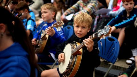 Boy looks at the camera, while playing the bango with a large group of young musicians. 