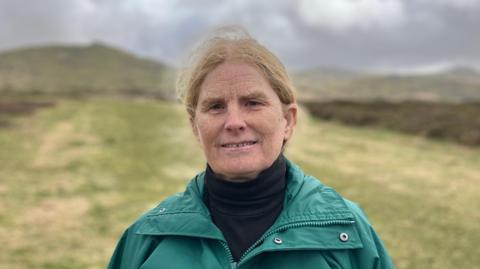 The image shows a woman wearing a green waterproof coat, standing on Dartmoor. There is a long strip of cut moorland - that acts as a firebreak - in the background. Either side of this is gorse, heather and other vegetation. The sky is grey and cloudy.