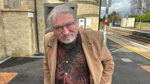 A man with glasses and a white goatie beard and windswept grey/white hair wearing a patterned shirt with a brown suede jacket over it, he is stood on a train platform.