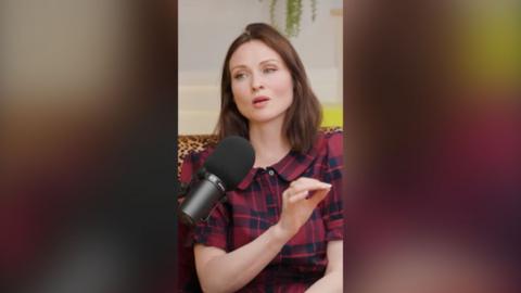 Sophie Ellis-Bextor sat with her hand raised mid-speech. She's wearing a Red and navy tartan top.
