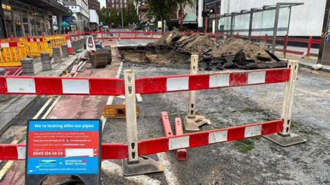 A construction site cordoned off with red and white barriers. A large hole has been excavated in the ground, surrounded by piles of dirt and debris. There is a sign which reads 'we're looking after our pipes'. In the background are more barriers and construction equipment.