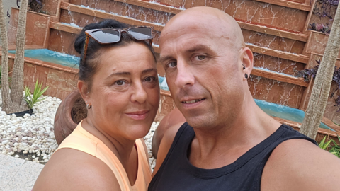 A woman with dark hair looks at the camera. She is wearing sunglasses on top of her head and an orange top. A man with a shaved head is next to her. He wears a black top and has two piercings in his ear.