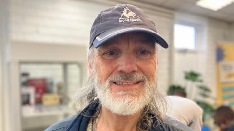 Stephen Smith, who has a white beard and long hair, smiles for a photo wearing a blue sports cap at one of the digital inclusion sessions. 