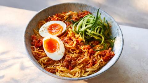 A grey bowl filled with noodles in an orange sauce, topped with a halved boiled egg