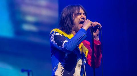 Bobby Gillespie has shoulder length dark hair and wears a colourful jacket of blue, white, yellow and pink. He sings into a microphone.