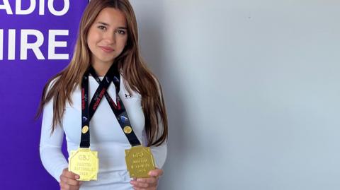Victoria standing in front of a banner. She has long brown hair and is wearing a long-sleeved white top and is holding her two gold medals