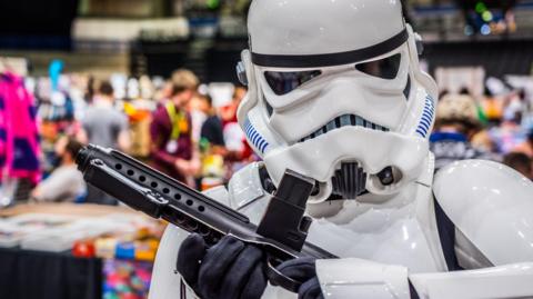 A person dressed as a stormtrooper standing in front of a crowd of people milling around stalls