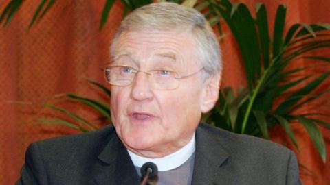 Rev Good giving a speech in front of a plant and an orange curtain. He is wearing a grey suit and a clerical collar