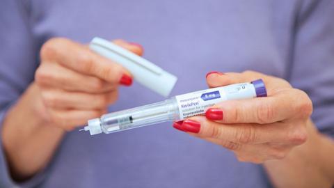 A woman's hands holding a weight loss jab pen, with the lid in her other hand - she has red nails and is wearing a lilac top 