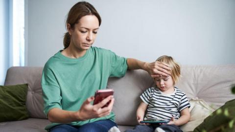 A woman on a sofa with tied back brown hair and a green top holding a pink phone in one hand and placing he other hand on the forehead of a small child with blonde hair and a black and white striped tshirt