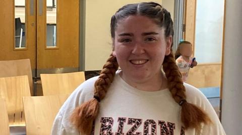 Aimee is standing in a vaccination clinic's waiting room. She has long brown hair in plaited bunches and is wearing a white jumper as she smiles for the camera.