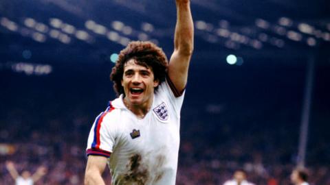 Kevin Keegan celebrates with his arm in the air after scoring a goal for England. He has short curly hair and is wearing a white England shirt with mud on the front.