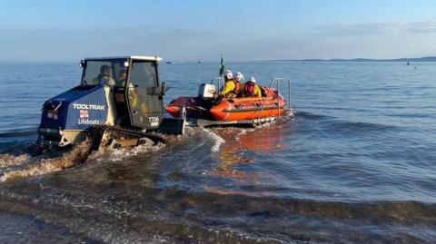 The RNLI inshore lifeboat is being pushed into the sea from the beach by a vehicle with tracks instead of tyres. The boat has three RNLI crew aboard, ready to launch into a calm sea with blue skies