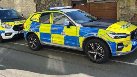 Two Guernsey Police vehicles. Both are painted yellow and blue with the word Police on the front of both. The second car, further to the left is a van, They are parked in front of a manky brown wall. 