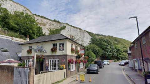 A old fashioned pub beside a narrow road. The pub and road are beneath white cliffs which are covered in trees and grass