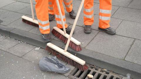 The legs of three people in hi-vis trousers who are sweeping up rubbish in the gutter on a road