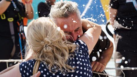 A man and a woman, wearing a navy dress with white spots, embrace. Confetti can be seen all around them.