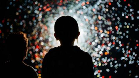 The silhouette of a young girl standings in front of a dark wall covered in brightly coloured lights.