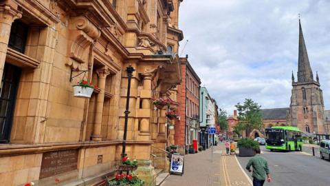 A quiet town street with a wide pavement runs past a large old stone building decorated with flowers. Across the road, a tall church with a pointed spire stands near a green bus and a few people walking.