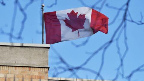 A Canadian flag flies above a grey building against the blue sky