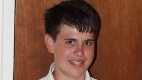 Jimmy Mizen smiling. It is a close up photo. He has dark hair and appears to be standing in front of an oak coloured door