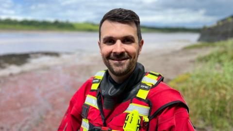 A river rescue worker wearing red and yellow safety uniform. He has short dark hair and facial hair and is standing on the banks of a river.