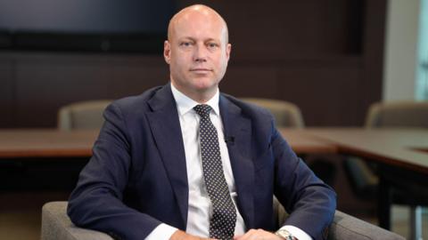 Dan Thomas sitting in a chair, wearing a blue suit, white shirt and pokadot tie.