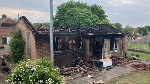 A small bungalow scorched black by fire. It's roof has collapsed and there is debris around the property.