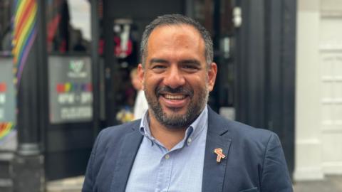 Manuel Guerrero Avina stands on a street, smiling at the camera, wearing a navy blazer and a blue shirt. He has a grey beard and grey hair.