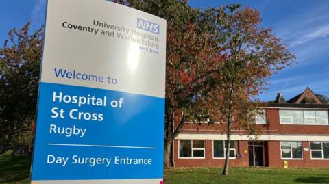 A blue and white hospital sign with the sun reflecting off the top. Tree and red brick building in the background. Sign reads, Hospital of St Cross, Rugby, with the NHS logo in the top right-hand corner. 